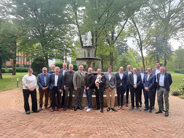 Eine Gruppe von 22 Personen steht um einen Brunnen in einem parkähnlichen Umfeld, umgeben von Bäumen und Grünflächen. Die Menschen lächeln und posieren für ein Gruppenfoto.