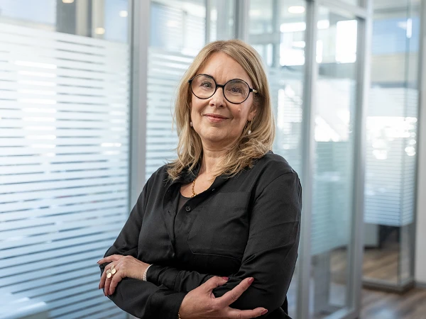 Eine lächelnde Frau mit langen blonden Haaren trägt eine schwarze Bluse und große Brillen. Sie steht in einem modernen Büro mit Glaswänden und kreuzt die Arme vor der Brust.