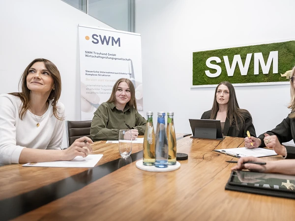 Vier Frauen sitzen an einem Konferenztisch in einem Besprechungsraum. Im Hintergrund sind ein Banner der SWM und ein Schild mit dem Schriftzug "SWM" zu sehen. Auf dem Tisch stehen Wasserflaschen und Gläser. Die Frauen schauen aufmerksam und sind mit Notizen und einem Tablet beschäftigt.