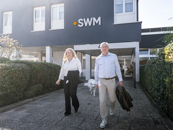 Eine Frau in einem weißen Hemd und ein Mann in einem weißen Hemd mit Jacke gehen vor einem modernen Gebäude mit dem Schriftzug "SWM", während ein weißer Hund neben ihnen läuft.
