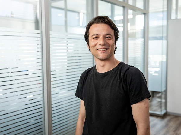 Ein lächelnder Mann mit kurzen, dunklen Haaren trägt ein schwarzes T-Shirt und steht in einem modernen Büro mit Glaswänden.