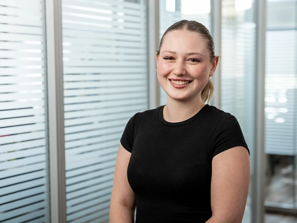 Eine lächelnde junge Frau mit einem dunklen T-Shirt steht in einem modernen, hellen Bürobereich mit Glaswänden im Hintergrund.