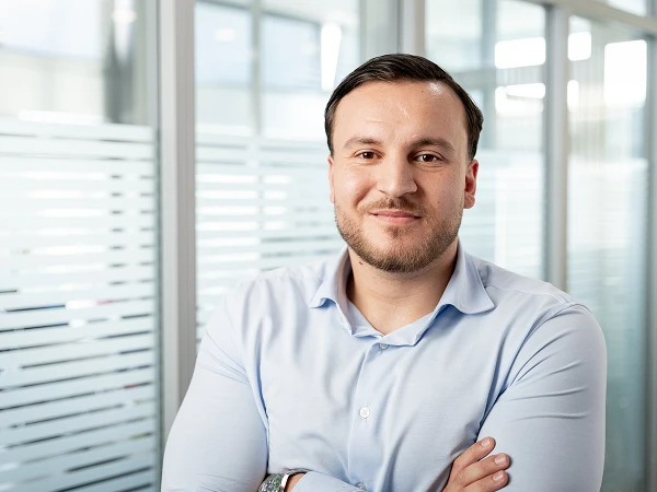 Ein Mann mit dunklen Haaren und leichtem Bart steht in einem modernen Büro, lächelt gut gelaunt und hat die Arme vor der Brust verschränkt. Im Hintergrund sind unscharf Fenster und Trennwände zu sehen.