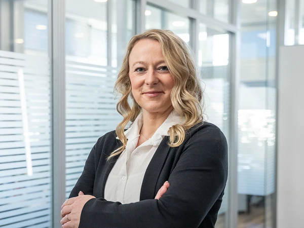 Eine Frau mit langen, welligen Haaren steht lächelnd mit verschränkten Armen in einem modernen Büro mit großen Fenstern und transparenten Trennwänden.