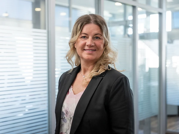 Eine Frau mit langen, gewellten blonden Haaren, die einen schwarzen Blazer und ein blumiges Oberteil trägt, steht in einem modernen Büroumfeld mit Glaswänden und viel natürlichem Licht.