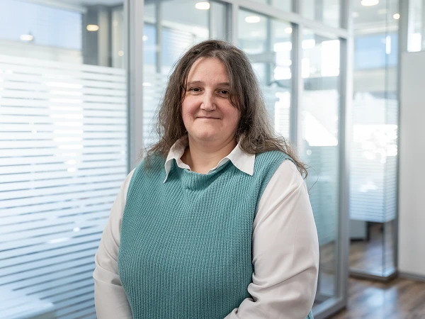 Eine Frau mit langen, lockigen Haaren steht in einem hellen Büro mit Glaswänden und modernen Möbeln. Sie trägt einen türkisfarbenen Pullover über einem weißen Hemd und lächelt freundlich in die Kamera.