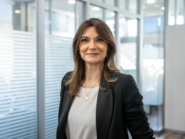 Eine Frau mit langen, braunen Haaren in einem schwarzen Blazer steht in einem modernen Büro. Sie lächelt und sieht in die Kamera, während hinter ihr Faltjalousien und Glaswände zu sehen sind.