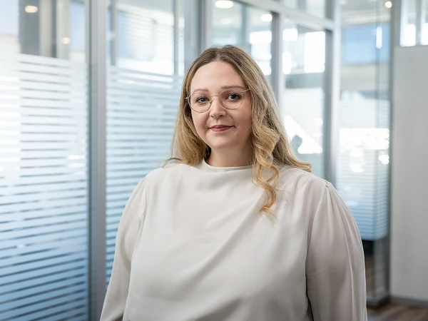 Eine Person mit langen, blonden, lockigen Haaren und einer hellen Bluse steht in einem modernen Büroumfeld mit Glaswänden und natürlichem Licht.