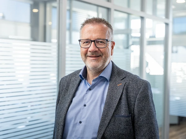 Ein Mann mittleren Alters mit Brille und grauem Bart steht in einem modernen Büroumfeld mit Glaswänden und verschiedenen Lichtquellen. Er trägt einen grauen Blazer und ein hellblaues Hemd, lächelt und sieht freundlich aus.