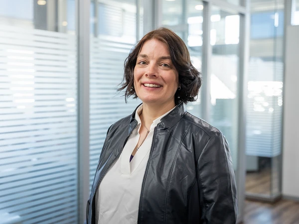 Eine lächelnde Frau mit dunklen, lockigen Haaren trägt eine schwarze Lederjacke und steht in einem modernen Büro mit Glaswänden.