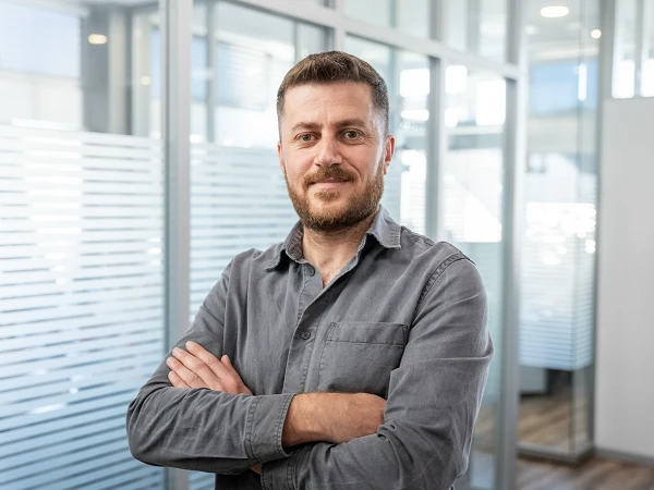 Ein Mann mit Bart in einem grauen Hemd steht mit verschränkten Armen in einem modernen Büro mit Glaswänden.