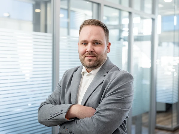 Ein Mann mit kurzen Haaren und einem grauen Sakko steht verschränkt in einem modernen Büro mit Glaswänden.