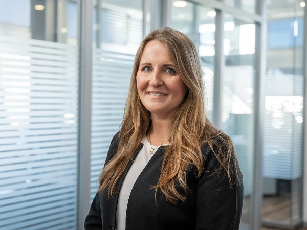 Eine lächelnde Frau mit langen, blonden Haaren trägt einen schwarzen Blazer und steht in einem modernen Büroraum mit Glaswänden und einem hellen, freundlichen Ambiente.