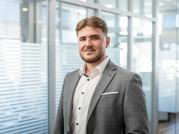 Junger Mann in einem hellgrauen Anzug und weißem Hemd, der in einem modernen, lichtdurchfluteten Büro steht und in die Kamera lächelt. Behind ihm sind verschwommene Glaswände und Büromöbel sichtbar.