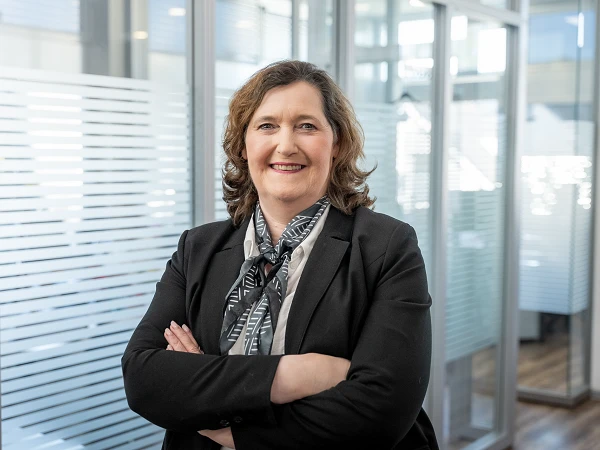 Eine beruflich gekleidete Frau mit lockigem, braunem Haar lächelt, während sie in einem modernen Büroraum mit großen Fenstern und Glaswänden steht. Sie hat die Arme verschränkt und trägt ein gestreiftes Halstuch.