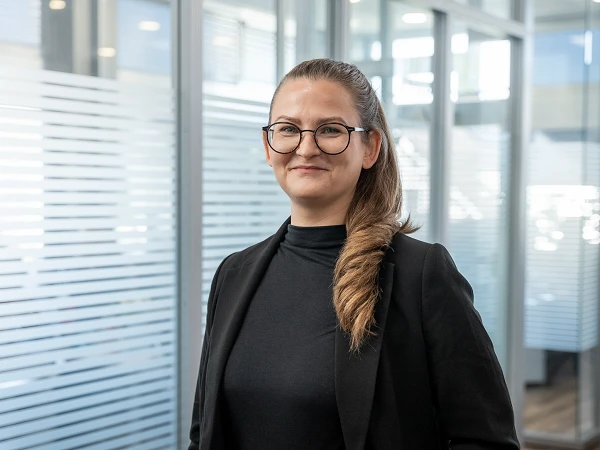 Eine Frau mit langen, gezopfteen Haaren und einer schwarzen Bluse sowie einem Blazer steht in einem modernen Büro mit Glaswänden und Holzboden. Sie lächelt und schaut in die Kamera.