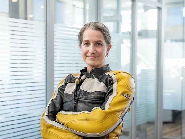 Eine Frau mit kurzen Haaren steht mit verschränkten Armen in einem modernen Büro. Sie trägt eine schwarze und gelbe Motorradjacke und blickt selbstbewusst in die Kamera. Im Hintergrund sind Glaswände sichtbar, die den Raum abtrennen.