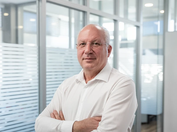 Ein Mann mit Brille und kurzen, grauen Haaren steht mit verschränkten Armen in einem modernen Büro mit Glaswänden. Er trägt ein weißes Hemd und lächelt leicht.