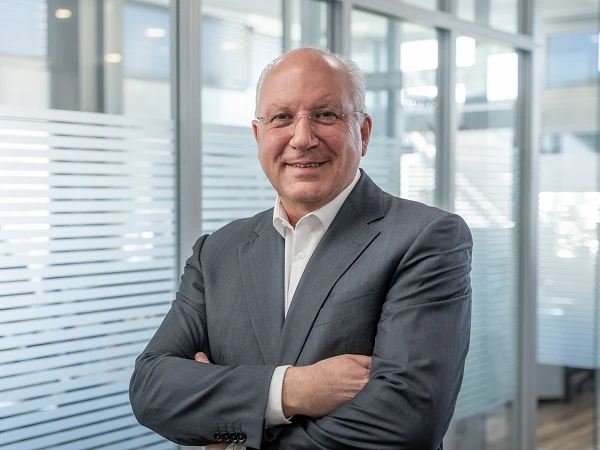 Ein lächelnder Mann in einem grauen Anzug steht mit verschränkten Armen vor einer Glaswand mit durchsichtigen Streifen in einem modernen Büro.