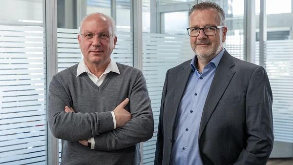 Zwei Männer stehen nebeneinander in einem modernen Büro mit transparenten Fensterscheiben. Der Mann links trägt einen grauen Pullover und hat die Arme verschränkt, während der Mann rechts einen dunklen Anzug und ein hellblaues Hemd trägt. Beide sehen direkt in die Kamera.