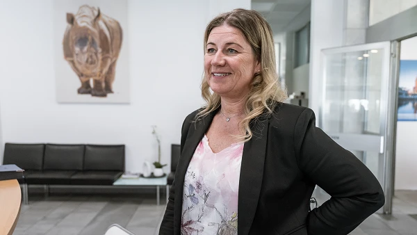 Eine lächelnde Frau im Business-Look steht in einem modernen Empfangsbereich, hinter ihr ist ein Bild eines Nashorns an der Wand zu sehen.