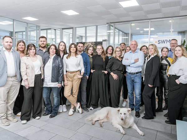 Eine Gruppe von 20 Personen steht in einem modernen Büro mit großen Fenstern, dabei ist ein Hund zu sehen. Die Menschen tragen unterschiedliche professionelle Kleidung und lächeln in die Kamera, einige stehen zusammen, während andere einzeln posieren.