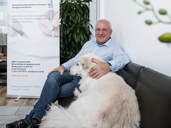 Ein Mann in einem hellblauen Hemd sitzt auf einem schwarzen Sofa in einem modernen Büro, während ein großer, flauschiger Hund entspannt neben ihm sitzt. Im Hintergrund ist ein Banner mit dem Logo „SWM“ und Informationen über die Firma zu sehen.
