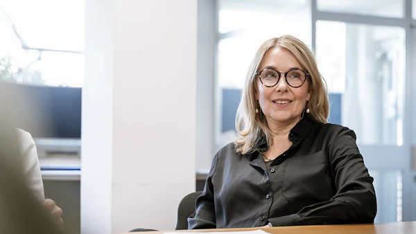 Eine Frau mit Brille sitzt an einem Tisch in einem modern eingerichteten Raum und lächelt, während sie an einem Gespräch teilnimmt. Auf dem Tisch liegt ein weißes Blatt Papier.