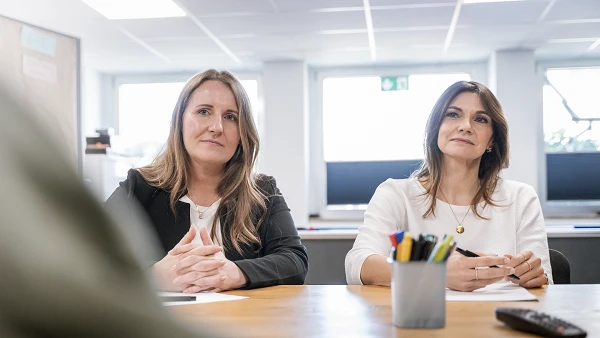 Zwei Frauen sitzen an einem Tisch in einem modernen Büro und hören aufmerksam zu, während jemand außerhalb des Bildes spricht. Vor ihnen steht ein Behälter mit verschiedenen Schreibutensilien.