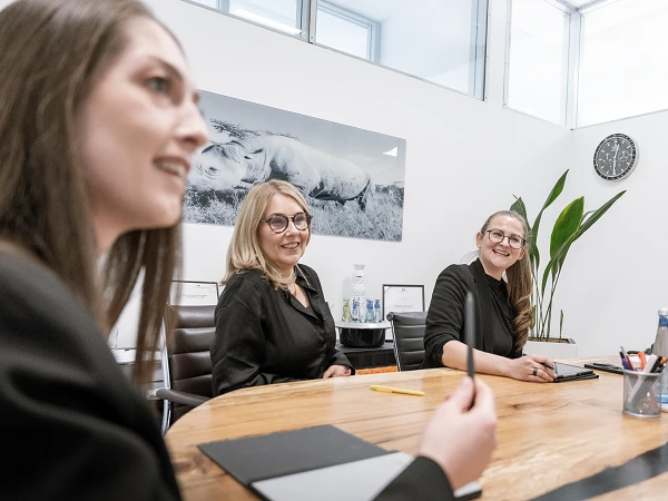 Drei Frauen in formeller Kleidung sitzen an einem Konferenztisch in einem modernen Besprechungsraum und diskutieren. Im Hintergrund ist ein Bild einer Landschaft sowie eine Uhr sichtbar. Auf dem Tisch stehen Schreibutensilien und Gläser mit Wasser.