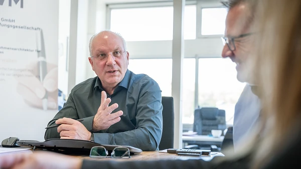 Ein älterer Mann mit Brille und einem grauen Hemd sitzt am Tisch und gestikuliert, während er spricht. Auf der linken Seite sind Teile eines weiteren Gesprächsteilnehmers und einige Büromaterialien sichtbar. Im Hintergrund sind Büromöbel und Tageslicht durch große Fenster zu sehen.