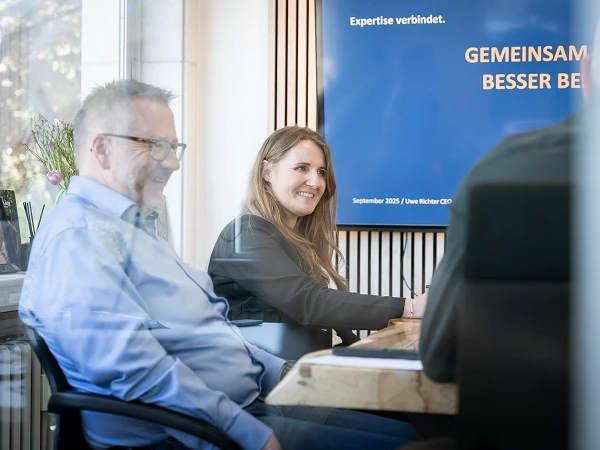 In einem Besprechungsraum sitzen ein Mann und eine Frau an einem Tisch und diskutieren. Die Frau lächelt, während der Mann einen freundlichen Gesichtsausdruck hat. Im Hintergrund ist ein Banner mit dem Text „Expertise verbindet“ und „GEMEINSAM BESSER BEI“ zu sehen.