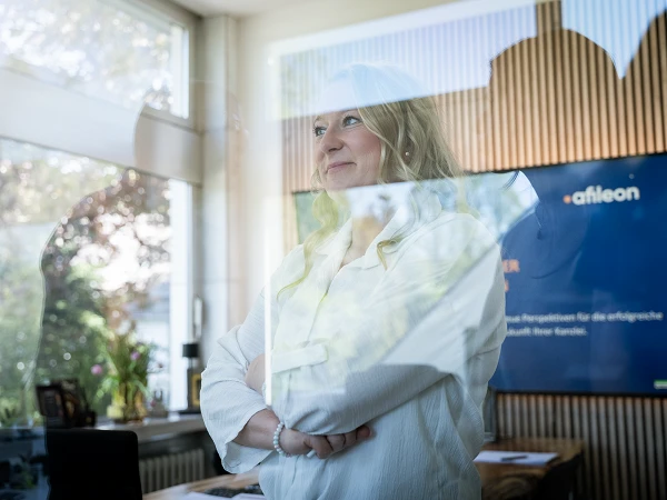Eine Person mit blonden, lockigen Haaren steht mit verschränkten Armen in einem modernen, hellen Raum mit großen Fenstern, hinter denen man Bäume sehen kann. Im Hintergrund ist ein Bildschirm zu sehen, auf dem Informationen über ein Unternehmen präsentiert werden.