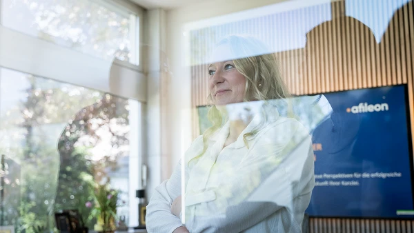Eine Person mit blonden, lockigen Haaren steht mit verschränkten Armen in einem modernen, hellen Raum mit großen Fenstern, hinter denen man Bäume sehen kann. Im Hintergrund ist ein Bildschirm zu sehen, auf dem Informationen über ein Unternehmen präsentiert werden.