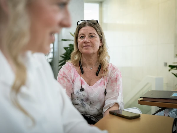 Eine blonde Frau mit einem blühenden Oberteil sitzt an einem Tisch, während eine andere Person im Vordergrund unscharf zu sehen ist. Im Hintergrund sind Pflanzen und helle Wände erkennbar.