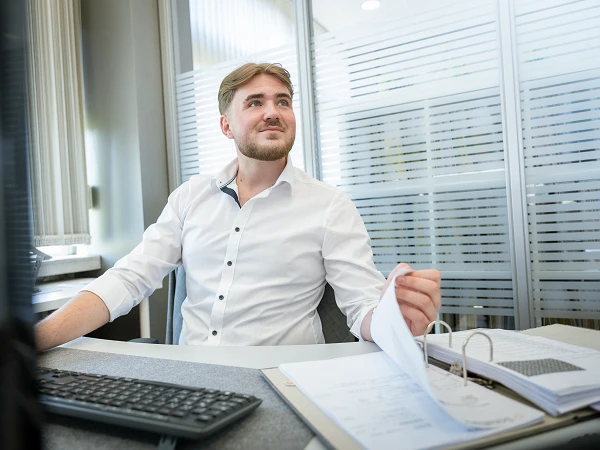 Ein Mann mit blonden Haaren sitzt an einem Schreibtisch, trägt ein weißes Hemd und schaut nachdenklich in die Ferne. Auf dem Tisch liegen mehrere Dokumente und ein Computer ist sichtbar.