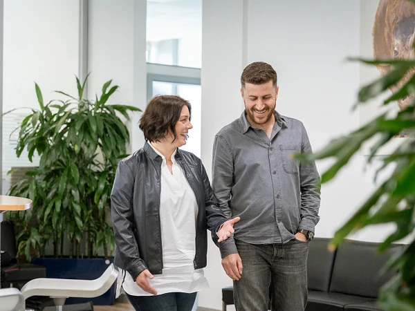 Ein Mann und eine Frau in lässiger Kleidung gehen in einem hellen, modernen Bürogebäude aufeinander zu und unterhalten sich, während sie an einer großen Pflanze vorbeigehen.