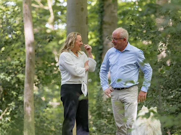 Ein Mann und eine Frau unterhalten sich im Wald, umgeben von grünen Bäumen. Die Frau trägt eine weiße Bluse und schwarze Hose, während der Mann ein hellblaues Hemd und beige Hosen anhat. Ein Hund ist ebenfalls im Bild sichtbar.