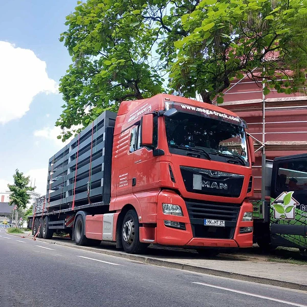 Ein roter Lkw von MAN parkt an einer Straße, neben einer grünen Fahrzeugen und teilweise verdeckt von einem Gebäude mit Gerüst. Im Hintergrund sind Bäume und ein klarer Himmel sichtbar.
