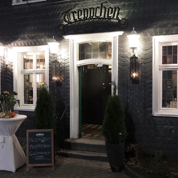 Der Eingang eines Restaurants mit dem Namen "Clemenschen" ist sichtbar, umgeben von eleganten, beleuchteten Wandleuchten. Vor der Tür stehen zwei kleine Pflanzen und ein Schild mit der Aufschrift "Restaurant", daneben ein Tisch mit Blumenarrangement.