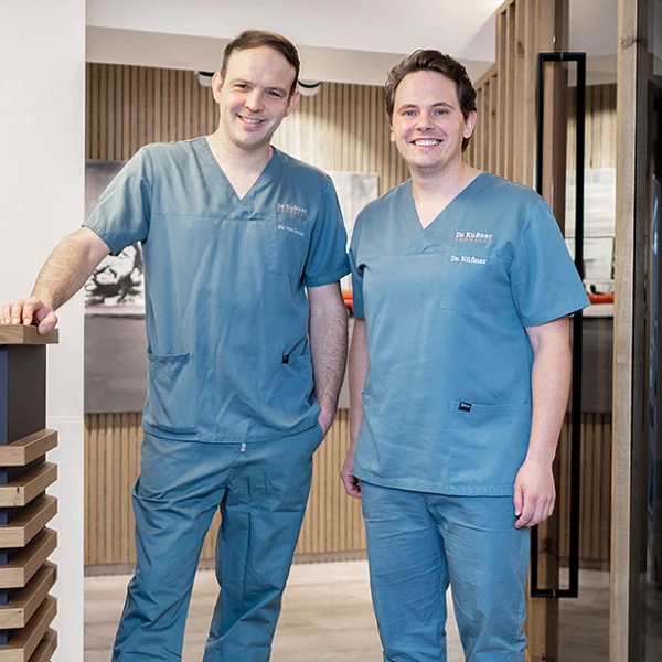 Zwei Zahnärzte in blauen Arbeitskleidungen stehen lächelnd am Empfangstresen einer modernen Zahnarztpraxis. Im Hintergrund ist das Praxisschild mit dem Namen "Dr. Kübner & Kollegen" sowie eine stilvolle Inneneinrichtung sichtbar.