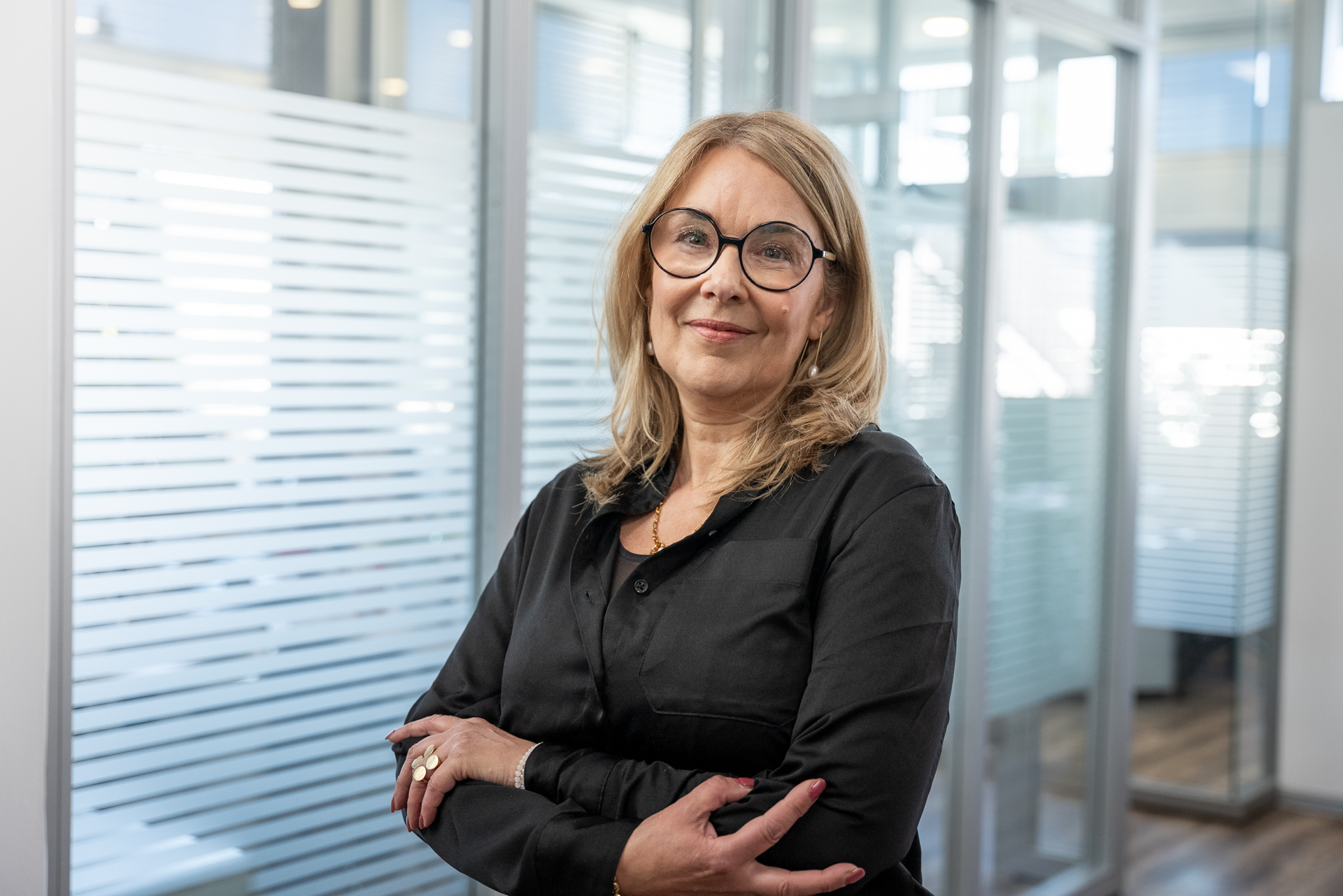 Eine lächelnde Frau mit langen blonden Haaren trägt eine schwarze Bluse und große Brillen. Sie steht in einem modernen Büro mit Glaswänden und kreuzt die Arme vor der Brust.