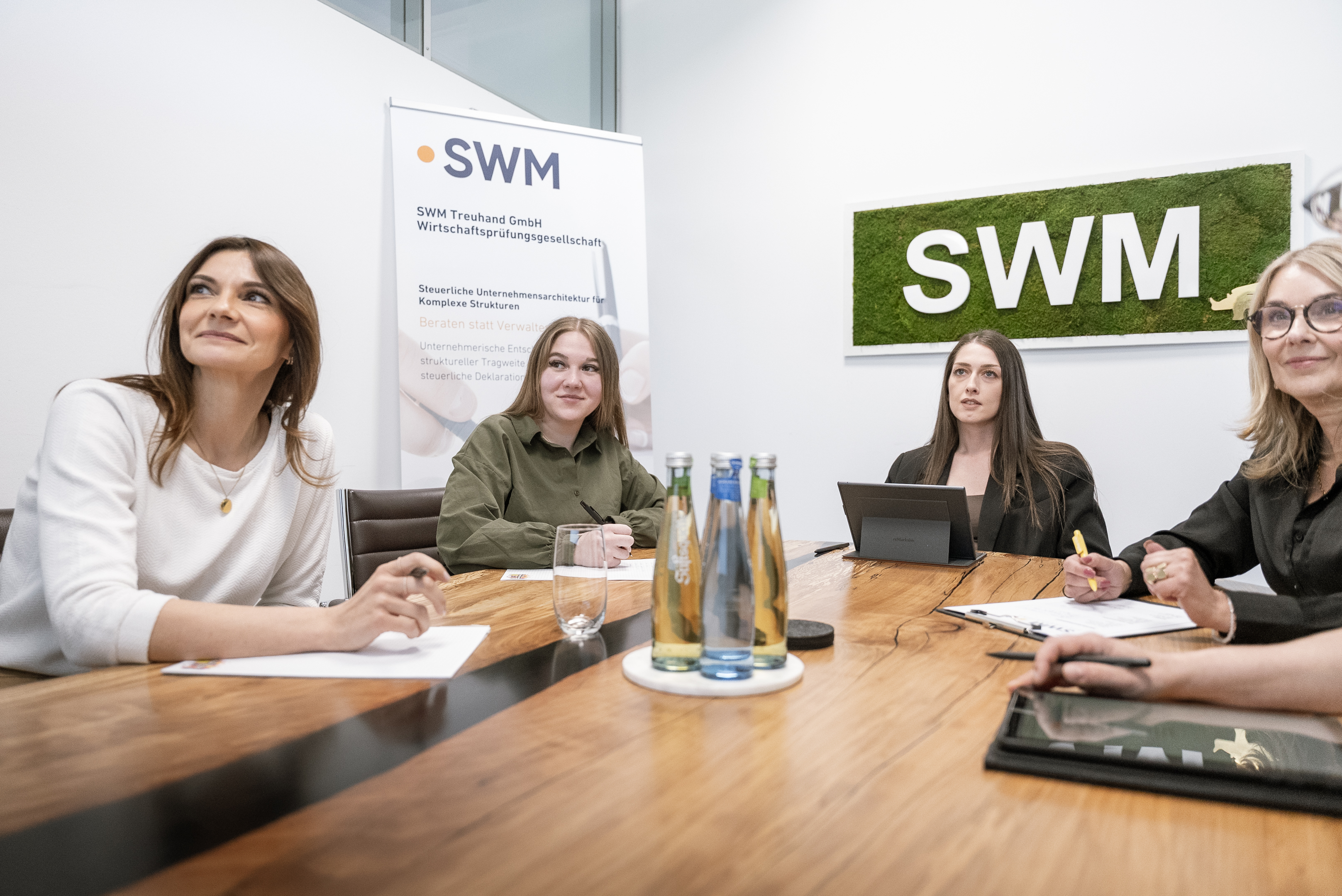Vier Frauen sitzen an einem Konferenztisch in einem Besprechungsraum. Im Hintergrund sind ein Banner der SWM und ein Schild mit dem Schriftzug "SWM" zu sehen. Auf dem Tisch stehen Wasserflaschen und Gläser. Die Frauen schauen aufmerksam und sind mit Notizen und einem Tablet beschäftigt.