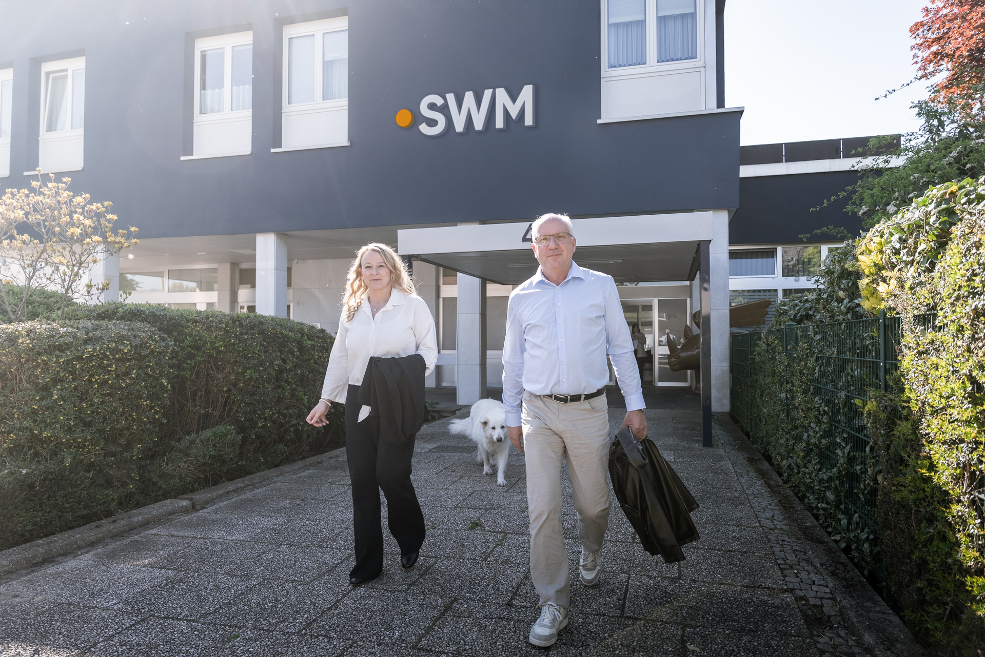 Eine Frau in einem weißen Hemd und ein Mann in einem weißen Hemd mit Jacke gehen vor einem modernen Gebäude mit dem Schriftzug "SWM", während ein weißer Hund neben ihnen läuft.