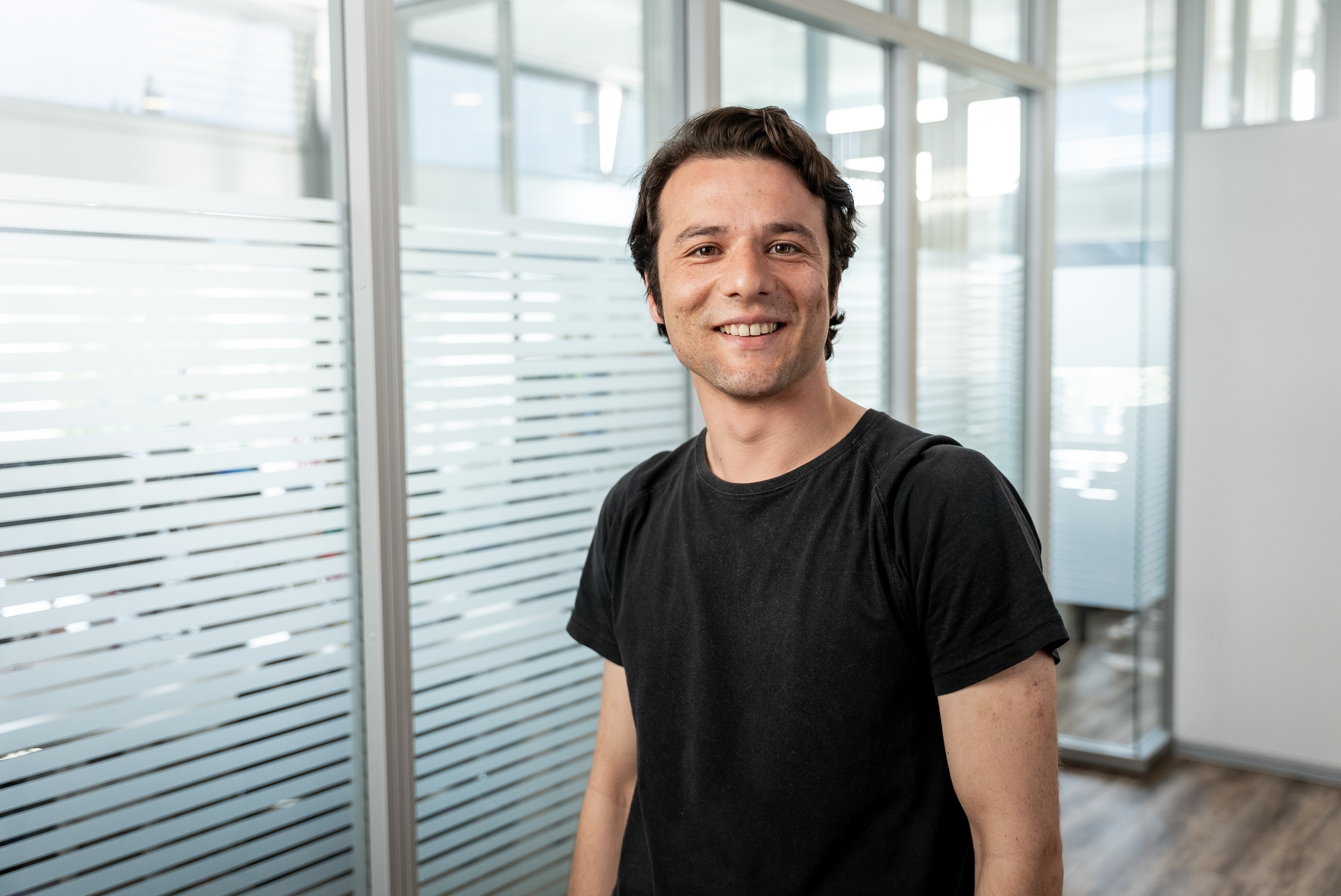 Ein lächelnder Mann mit kurzen, dunklen Haaren trägt ein schwarzes T-Shirt und steht in einem modernen Büro mit Glaswänden.