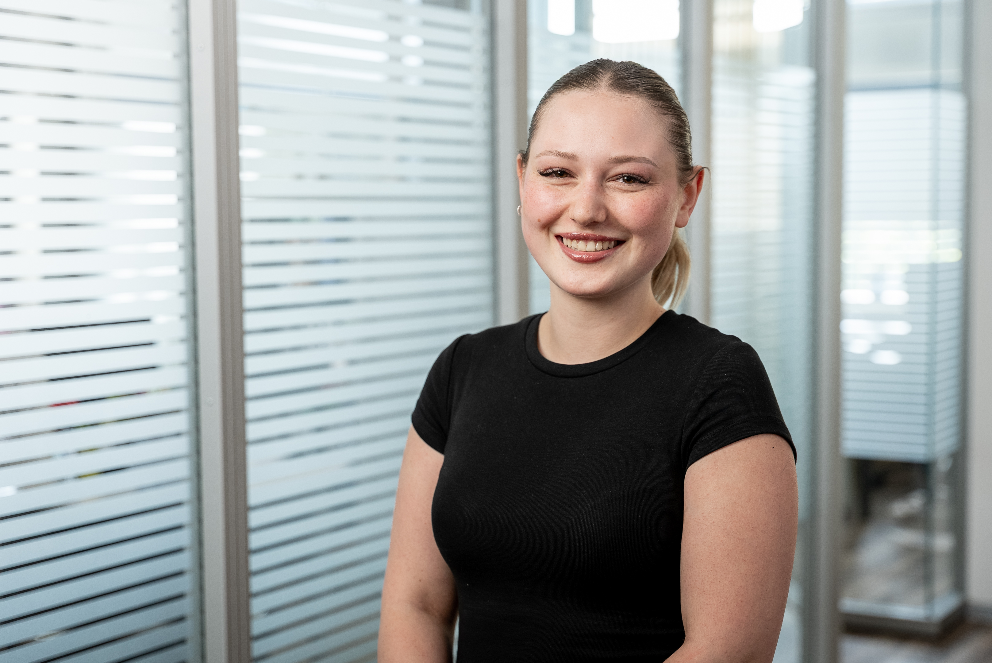 Eine lächelnde junge Frau mit einem dunklen T-Shirt steht in einem modernen, hellen Bürobereich mit Glaswänden im Hintergrund.