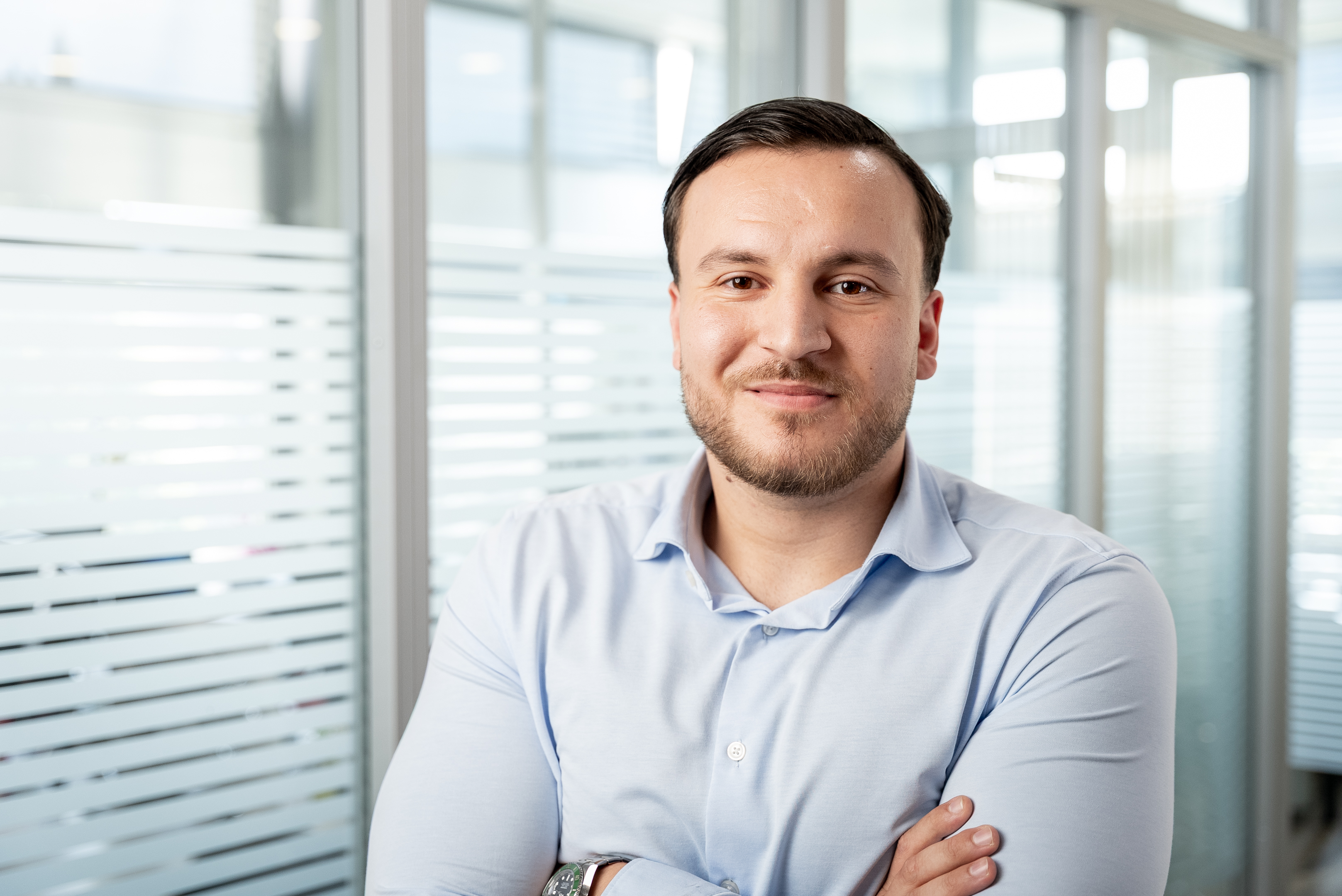 Ein Mann mit dunklen Haaren und leichtem Bart steht in einem modernen Büro, lächelt gut gelaunt und hat die Arme vor der Brust verschränkt. Im Hintergrund sind unscharf Fenster und Trennwände zu sehen.