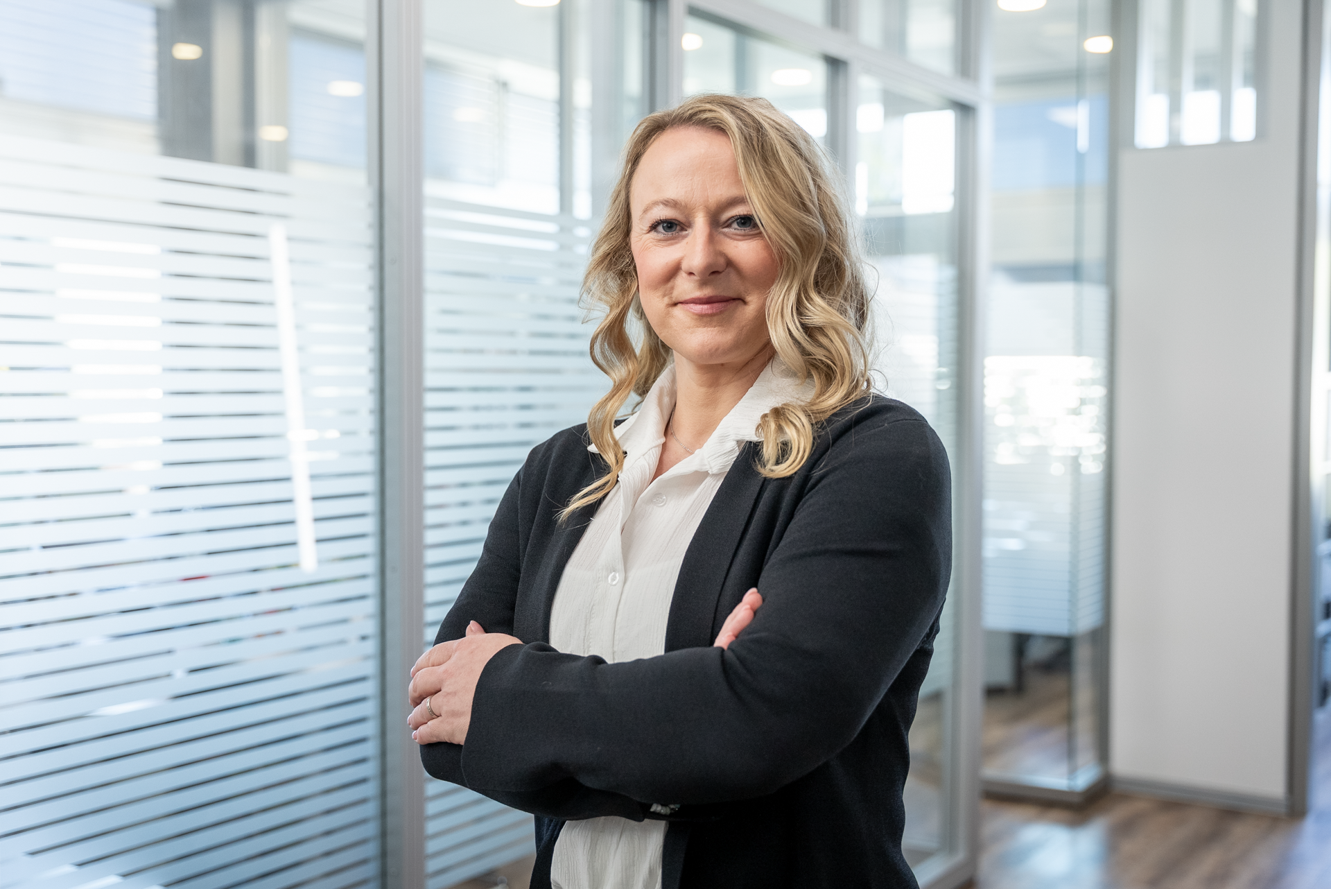 Eine Frau mit langen, welligen Haaren steht lächelnd mit verschränkten Armen in einem modernen Büro mit großen Fenstern und transparenten Trennwänden.