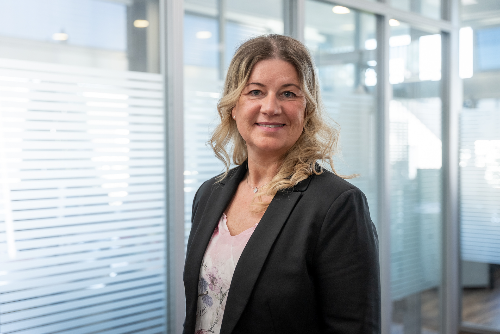 Eine Frau mit langen, gewellten blonden Haaren, die einen schwarzen Blazer und ein blumiges Oberteil trägt, steht in einem modernen Büroumfeld mit Glaswänden und viel natürlichem Licht.