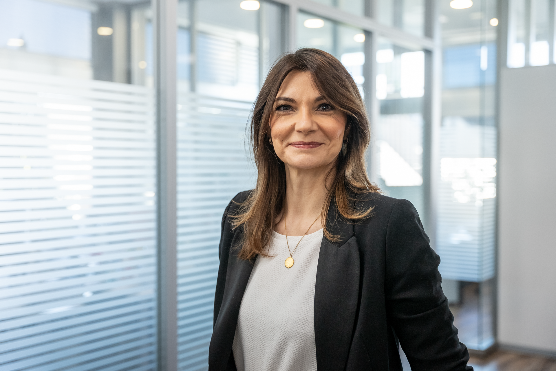 Eine Frau mit langen, braunen Haaren in einem schwarzen Blazer steht in einem modernen Büro. Sie lächelt und sieht in die Kamera, während hinter ihr Faltjalousien und Glaswände zu sehen sind.